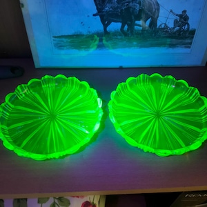 Peut inclure: Deux assiettes en verre vert vif assorties, à bords festonnés. Les assiettes présentent un motif radial partant du centre. Les assiettes sont posées sur une surface en bois. Une image encadrée est en arrière-plan.
