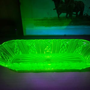 May include: A glowing green glass dish with a rectangular shape and intricate geometric patterns. The dish has a textured surface and a sunburst design at the base. The dish is likely vintage and made of uranium glass.