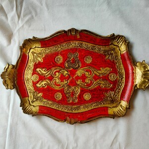 Pode incluir: Uma bandeja decorativa retangular com base vermelha e acabamento dourado. A bandeja apresenta volutas e alças douradas ornamentadas. A bandeja tem aproximadamente 46 cm de comprimento e 30 cm de largura.