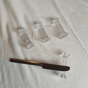 May include: Four clear glass knife rests with a twisted column design. A brown-handled knife with a dark blade rests on one. The knife rests are on a white tablecloth.
