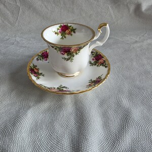Könnte beinhalten: Ein weißes Porzellan-Tassenset mit Untertasse, verziert mit einem zarten Blumenmuster aus rosa, roten und gelben Rosen mit grünen Blättern. Der Rand der Tasse und der Untertasse ist mit einem dünnen, polierten Goldrand versehen, und die Tasse hat einen Henkel.