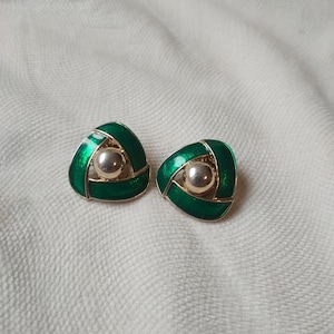 Puede incluir: Un par de pendientes de botón. Cada pendiente presenta un diseño triangular de esmalte verde esmeralda con una bola plateada en el centro. Los pendientes están sobre un fondo blanco texturizado.