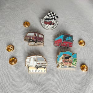 Puede incluir: Cinco pines de esmalte con diferentes vehículos. Los pines están sobre un fondo de tela blanca. Los pines representan un autobús rojo, una furgoneta blanca con el texto "Server", una furgoneta blanca con el texto "Exporter", un camión rojo con un fondo azul y blanco, y un camión rojo con una bandera a cuadros blanca y el texto "Mercedes-Benz Competition".