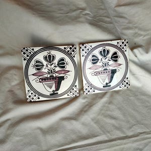 May include: Two square ceramic tiles with a floral design. Each tile features a black and white circular border with a central image of a vase holding flowers in black, white, and mauve. The tiles are set against a neutral background.