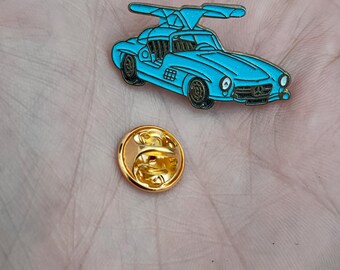 May include: A blue enamel pin of a vintage car with a propeller on top. The pin is on a hand and has a gold back.
