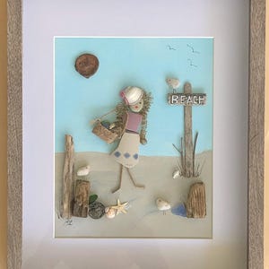May include: Framed artwork depicting a person walking on a beach, carrying a basket. The artwork is made of natural materials, with a blue sky background and a sign that says "BEACH". The artwork is framed in a gray wooden frame.