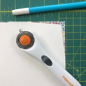 Puede incluir: Un cortador rotatorio blanco con un botón naranja sobre una estera de corte verde. El cortador está sobre un trozo de papel blanco con un agujero.