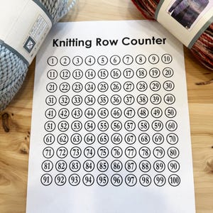 Könnte beinhalten: Ein weißes Blatt Papier mit dem Text "Knitting Row Counter" in Schwarz oben gedruckt. Das Papier ist in ein Raster aus 10 Spalten und 10 Zeilen unterteilt, wobei jede Zelle einen schwarzen Kreis mit einer weißen Zahl darin enthält. Die Zahlen reichen von 1 bis 100.