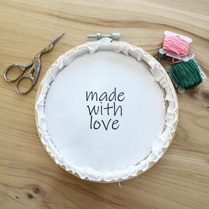 May include: A white embroidery hoop with the text "made with love" on the fabric inside. The hoop is on a wooden surface with pink and green embroidery floss.