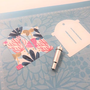 Puede incluir: Una plantilla de papel blanco con tres ranuras y un agujero en la parte superior, una plantilla de papel con estampado floral con tres ranuras y una herramienta plateada para perforar agujeros en el papel.