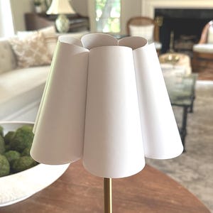 Puede incluir: Una pequeña lámpara de papel blanco con una pantalla única en forma de pétalo. La lámpara tiene una base cuadrada dorada y un tallo delgado. Está sobre una mesa de madera oscura, con un salón borroso en el fondo.