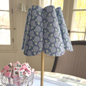 May include: A decorative table lamp with a blue floral patterned lampshade. The lamp has a gold-colored base and stem. A glass bowl filled with pink and white Mahjong tiles sits nearby. The lamp is on a light blue tablecloth.