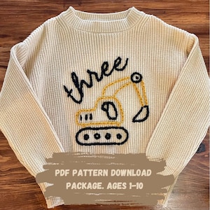 Peut inclure: Un pull en tricot beige avec un excavateur brodé en noir et jaune et le mot "three" en broderie noire. Le pull est pour les âges 1-10. Téléchargement du modèle PDF.