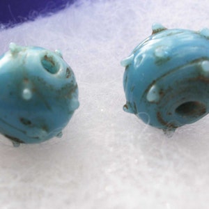 May include: Two turquoise blue glass beads with a rough, textured surface and small bumps. The beads have a hole in the center for stringing.