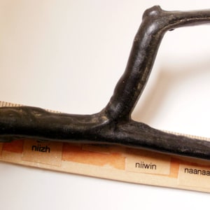 May include: A black wooden stick bent into a U-shape. The stick is resting on a ruler with the words "bezhig", "niizh", "niiwon", and "naanaan" printed on it.