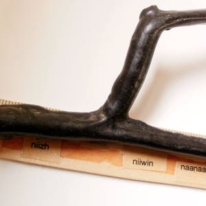 May include: A black wooden stick bent into a U-shape. The stick is resting on a ruler with the words "bezhig", "niizh", "niiwon", and "naanaan" printed on it.