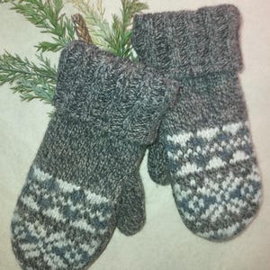 May include: A pair of gray knitted mittens with a folded cuff. The mittens feature a geometric pattern in white and blue on the lower portion. The mittens are displayed on a light background with green sprigs.