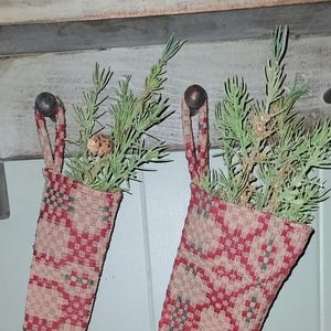 Puede incluir: Dos calcetines navideños con patrones geométricos rojos y verdes, llenos de ramitas verdes y pequeñas piñas. Los calcetines cuelgan de ganchos de metal marrón oscuro sobre una superficie de madera clara.