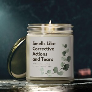 May include: A lit candle in a clear glass jar with a silver lid. The label reads "Smells Like Corrective Actions and Tears" with a botanical illustration. The candle is a natural wax blend and is vegan.