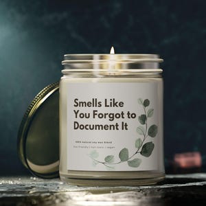 May include: A clear glass jar candle with a silver lid, labeled "Smells Like You Forgot to Document It." The label features green eucalyptus leaves. The candle is lit, with a small flame. The candle is on a dark surface.