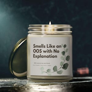 Könnte beinhalten: Eine brennende Kerze in einem durchsichtigen Glas mit einem silbernen Deckel. Auf dem Etikett steht "Smells Like an OOS with No Explanation" mit einem botanischen Design. Die Kerze ist eine natürliche Sojawachsmischung, umweltfreundlich und vegan.