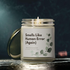 May include: A lit soy wax candle in a clear glass jar with a silver lid. The label reads "Smells Like Human Error (Again)" with a botanical design. The candle is on a dark surface.
