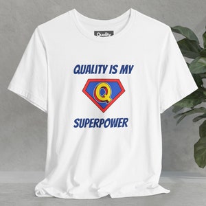 May include: White t-shirt with the text "QUALITY IS MY SUPERPOWER" in blue, and a superhero-style emblem with a yellow "Q" inside a red and blue diamond. The shirt has a "Quality" tag.