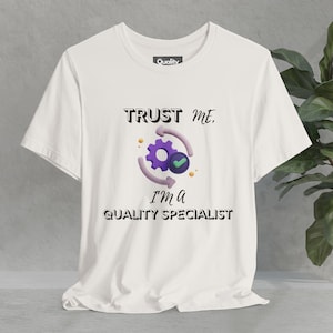 Op de afbeelding: Wit T-shirt met de tekst "TRUST ME, I'M A QUALITY SPECIALIST" en een paars tandwielpictogram met een groen vinkje erin.