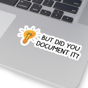May include: A white sticker with the text "BUT DID YOU DOCUMENT IT?" and a yellow lightbulb graphic. The sticker is placed on a silver laptop, near the keyboard.
