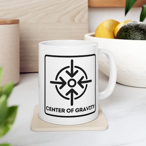 Puede incluir: Taza de cerámica blanca con un gráfico negro de flechas que apuntan hacia un círculo central, con el texto "CENTER OF GRAVITY" debajo. La taza está sobre un posavasos beige. La taza tiene un asa blanca.