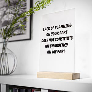 Op de afbeelding: Een helder acryl bord met de tekst "LACK OF PLANNING ON YOUR PART DOES NOT CONSTITUTE AN EMERGENCY ON MY PART" in zwart, geplaatst op een lichte houten basis. Het bord staat op een witte plank met een vaas met groen.