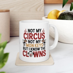 Puede incluir: Taza de cerámica blanca con el texto "NOT MY CIRCUS NOT MY MONKEYS BUT I KNOW THE CLOWNS" en rojo y negro. La taza presenta gráficos de carpa de circo y monos. La taza está sobre un posavasos beige.