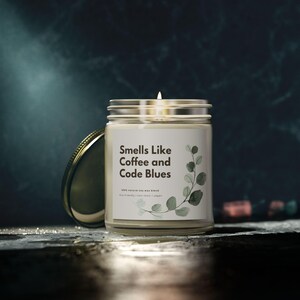 Peut inclure: Une bougie allumée dans un pot en verre transparent avec un couvercle argenté. L'étiquette indique "Smells Like Coffee and Code Blues" avec une illustration botanique. La bougie est sur une surface sombre avec un arrière-plan flou.