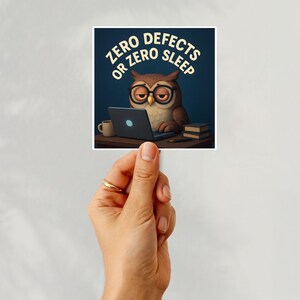 May include: A sticker featuring a cartoon owl wearing glasses, working on a laptop. The text reads "ZERO DEFECTS OR ZERO SLEEP" in a circular design. A coffee mug and books are on the desk.
