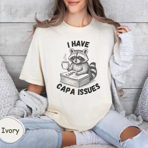 May include: Ivory-coloured t-shirt with a raccoon illustration holding a coffee cup and sitting on books. The text "I HAVE CAPA ISSUES" is printed in black. The shirt has a relaxed fit.