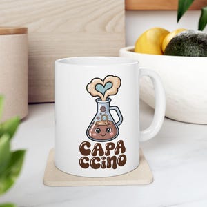 May include: White ceramic mug featuring a cartoon illustration of a beaker with a face, steam shaped like a heart, and the text "CAPACCINO" in brown. The mug is on a beige coaster.