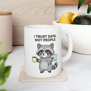 Può includere: Tazza in ceramica bianca con un procione dei cartoni animati che tiene in mano una tazza di caffè e un grafico. La tazza riporta la scritta "I TRUST DATA. NOT PEOPLE." Il procione indossa un distintivo QA. La tazza è appoggiata su un sottobicchiere beige.