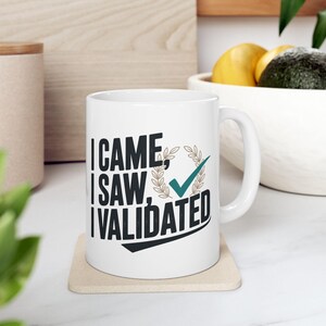 Può includere: Tazza in ceramica bianca con la scritta "I CAME, I SAW, I VALIDATED" in nero, con un segno di spunta verde acqua e un design a corona d'alloro. La tazza è appoggiata su un sottobicchiere beige.