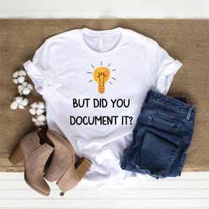 以下が含まれることがあります： 電球のグラフィックと「But did you document it?」というテキストが書かれた白いTシャツ