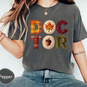 May include: Dark grey t-shirt featuring the word "DOCTOR" in a patchwork design. The letters incorporate autumnal motifs like a maple leaf and acorn, with plaid and textured fabrics, creating a seasonal aesthetic.