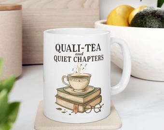 May include: White ceramic mug with the text "QUALI-TEA and QUIET CHAPTERS" and a graphic of a teacup on a stack of books. The teacup is beige with a brown liquid and steam rising. A pair of glasses is at the bottom.