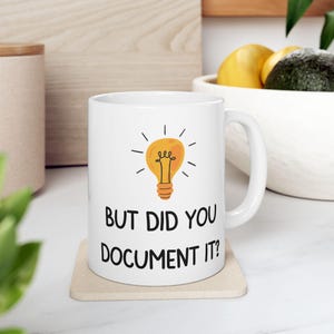 Op de afbeelding: Witte keramische mok met een zwarte gloeilamp afbeelding en de tekst "But did you document it?" erop gedrukt.