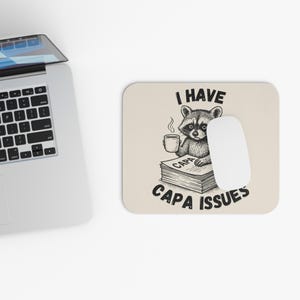 May include: A beige rectangular mouse mat featuring a raccoon illustration holding a coffee cup, with the text "I HAVE CAPA ISSUES". A white mouse and a laptop are also visible in the image.