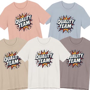 Op de afbeelding: Een collectie van vijf T-shirts in verschillende kleuren: lichtroze, lichtblauw, beige, wit en grijs. Elk shirt heeft de tekst "QUALITY TEAM" in een stripboekstijl ontwerp met een gele en rode explosie.