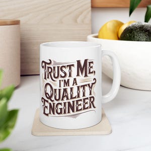 Op de afbeelding: Witte keramische mok met de tekst "Trust Me, I'm a Quality Engineer" in bruine letters. De mok staat op een beige onderzetter. De mok heeft een wit handvat.