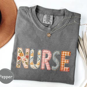 May include: A gray Comfort Colors t-shirt with the word "NURSE" appliqued across the front. The letters are made of various patterned fabrics, including floral, patchwork, and gingham. A silver necklace with a pendant is visible.