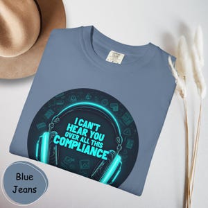 Könnte beinhalten: Ein blaues T-Shirt mit einer Grafik von Kopfhörern in Neonblau und dem Text "I CAN'T HEAR YOU OVER ALL THIS COMPLIANCE". Das Shirt hat eine einfarbige blaue Farbe und ein Etikett oben rechts.