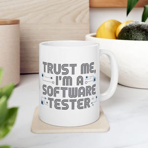 Op de afbeelding: Witte keramische mok met de tekst "TRUST ME, I'M A SOFTWARE TESTER" in zwart en blauw. De mok heeft een gebogen oor en staat op een beige onderzetter. De mok is een standaard formaat.