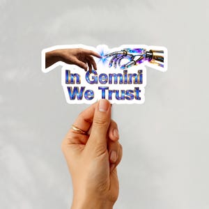 Op de afbeelding: Een sticker die door een hand wordt vastgehouden, met de tekst "In Gemini We Trust" in blauw en goud. Het ontwerp toont een menselijke hand die een robotachtige hand aanraakt met een vonk ertussen. De achtergrond is effen wit.