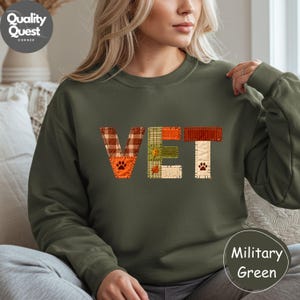Puede incluir: Sudadera verde militar con la palabra "VET" en letras de patchwork. Las letras están hechas de varios patrones, incluyendo cuadros y colores lisos, con huellas de patas. La sudadera es de un estilo cómodo e informal.